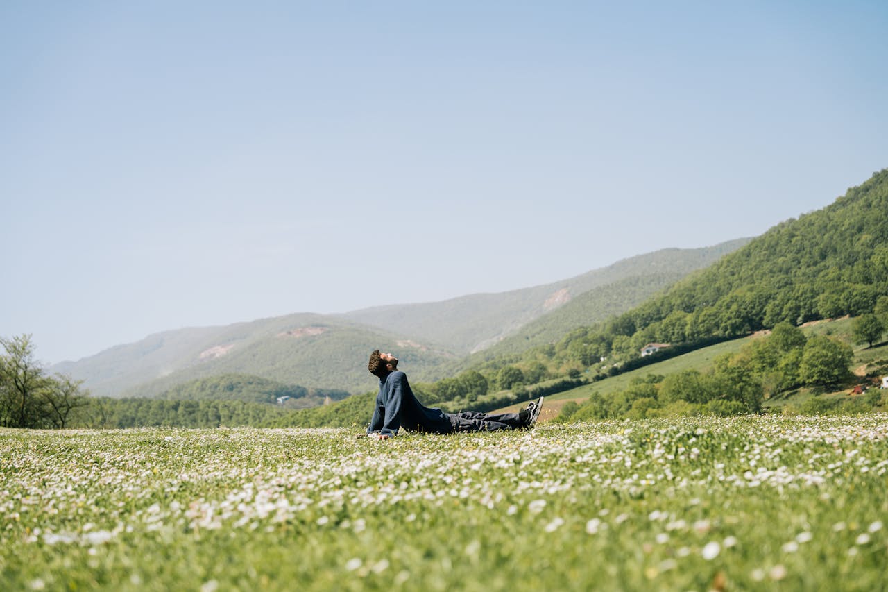A man relaxes on a daisy-covered field with scenic green hills and a clear sky.