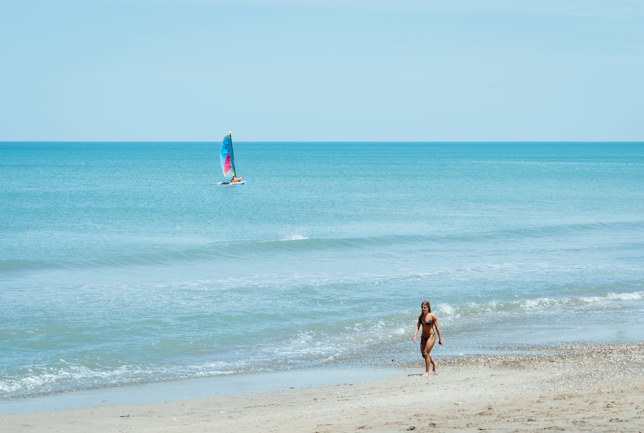 A woman walks along Mar del Plata beach with a sailboat in the distance on a sunny day.